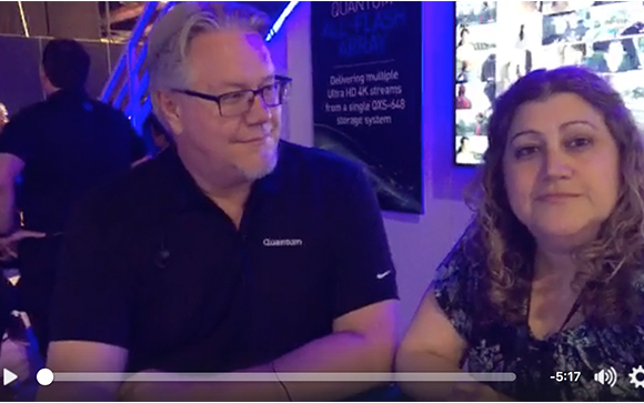 A man with glasses and a woman with curly hair sitting in front of a blue illuminated sign.