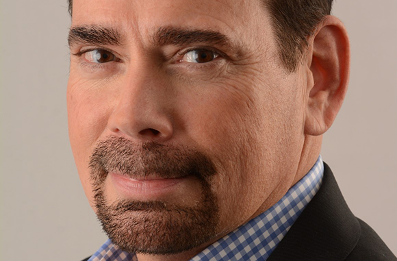 Close-up portrait of a man with dark hair, brown eyes, and a groomed goatee wearing a blue checkered shirt.