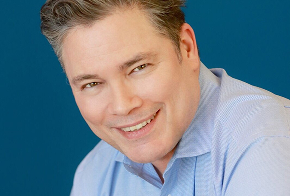 A smiling man with short brown hair wearing a light blue collared shirt against a solid blue background.