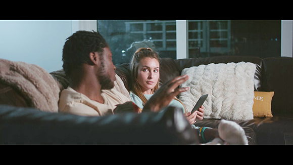 A man and a woman sit together on a dark brown couch in a dimly lit room.