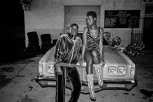 A man in a velvet tracksuit and a woman in a zebra print top sit on a vintage car outside a garage.