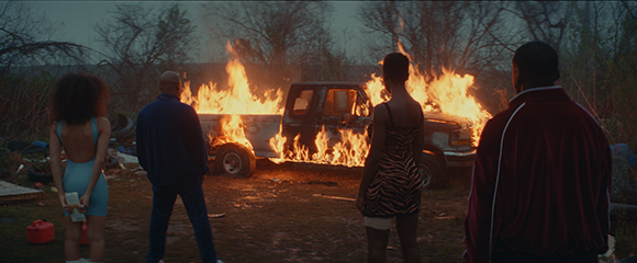 Four people stand with their backs to the camera, watching a pickup truck engulfed in flames at dusk.