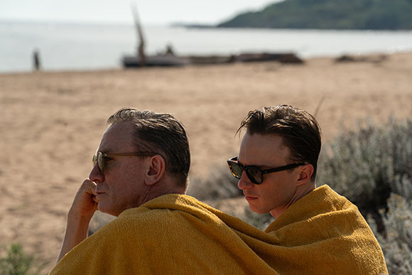 Two men wearing sunglasses and wrapped in yellow towels sit on a sandy beach.