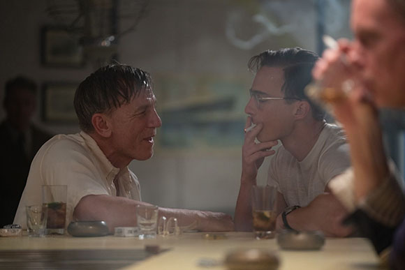 Two men sit at a table in a dimly lit room, one smoking a cigarette while looking at the other.