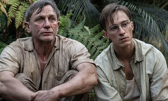 Two men in dirty, light-colored shirts sit side-by-side amidst dense green tropical foliage.