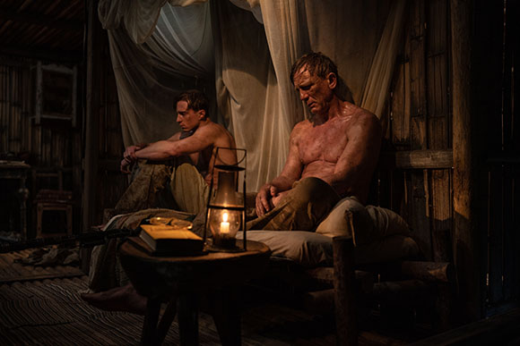 Two shirtless men sit in a dark, rustic room illuminated by a small lantern on a table.