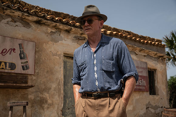 A man wearing a fedora, sunglasses, and a blue button-down shirt stands before a rustic, aged building.