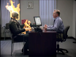 A man in a blue shirt sits at a desk facing a person with a flaming skull and hands.