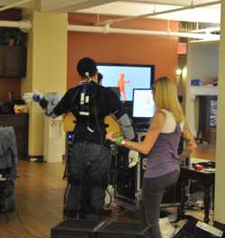 A person in a black motion capture suit stands before a screen as a woman in a purple tank top watches.