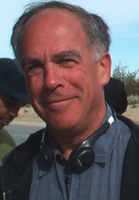 A middle-aged man with short grey hair smiling, wearing a striped shirt and headphones around his neck.