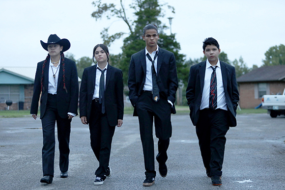Four teenagers in black suits and ties walking forward on an asphalt surface outdoors.
