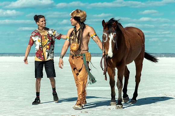 Two men walk across a vast white salt flat next to a brown horse under a blue sky.