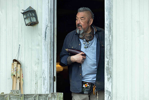 An older man with a grey beard and goggles around his neck stands in a doorway holding an eggplant.
