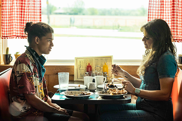 A young man and a woman sit at a small table in a diner, eating food near a window.