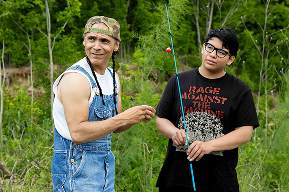 A man in denim overalls and a person in a black t-shirt stand in a grassy field holding a blue fishing rod.