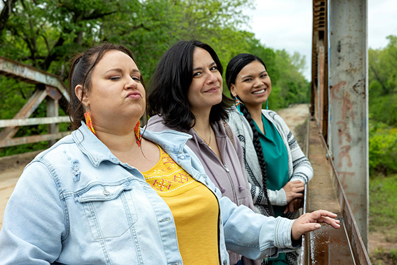 Three women smiling and posing together on a metal bridge outdoors with green trees in the background.