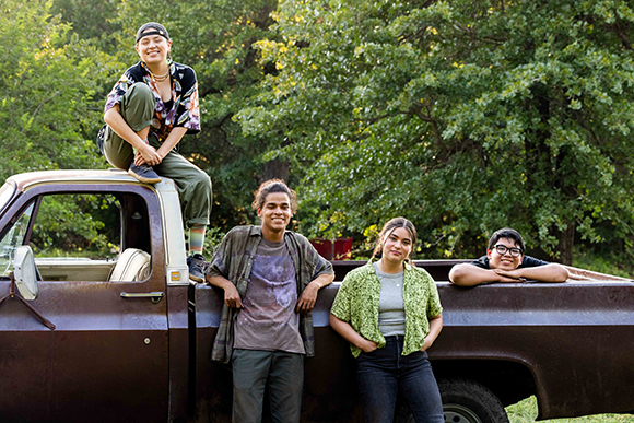 Four smiling young people pose with an old brown pickup truck in a lush, green wooded setting.