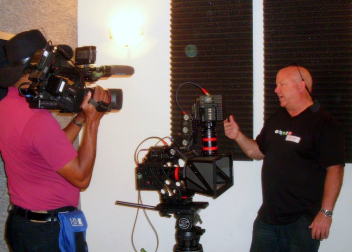 A cameraman in a pink shirt and black hat operates a professional video camera. A man in a black t-shirt gestures towards a R