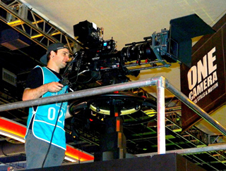 A man in a blue jersey operates a large professional film camera mounted on a pedestal.