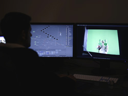 A person sits in a dark room facing two computer monitors displaying video editing software and a green screen clip.