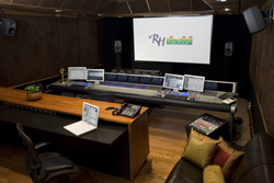 A professional audio mixing studio with a large console, multiple monitors, and a projection screen.