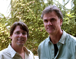 Two men standing side by side outdoors in front of a wall of green climbing plants.