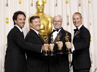 Four men in black tuxedos smiling and holding gold Oscar trophies in front of a large gold Oscar statue.