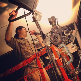 A man on a red ladder adjusts a large professional film camera and lighting rig.