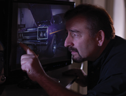 A man in a dark shirt points at a photo of a purple car displayed on a computer screen.