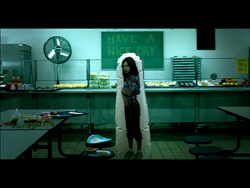 A person dressed as a hot dog stands in a cafeteria under a sign that reads 'HAVE A NICE DAY'.