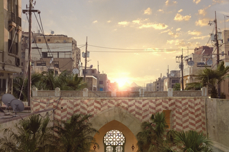 Sun setting behind buildings and utility poles in a city with palm trees and stone walls.