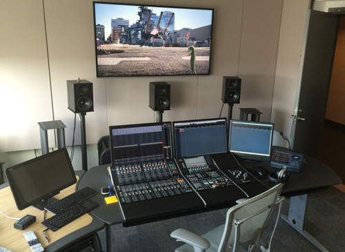 A large desk with multiple computer monitors, a digital mixing console, studio monitors, and a large wall-mounted screen.