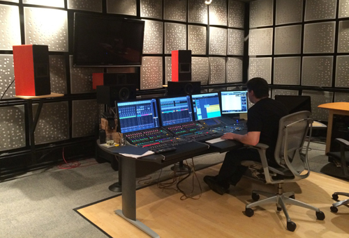 A person sits at a desk with multiple computer monitors and a large audio mixing console in a studio.