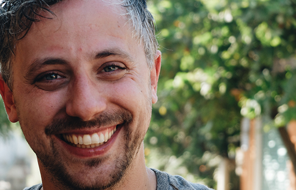 A close-up shot of a man with short, dark, wavy hair and a light beard, smiling broadly.
