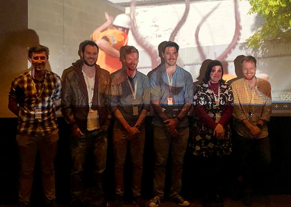 Six people stand in a row in front of a large projection of a person in a safety vest.