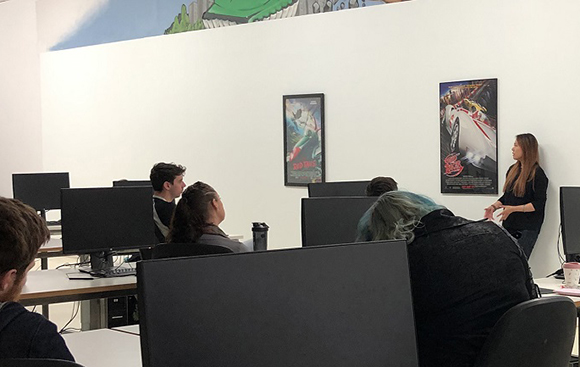 Students sit at computer desks in a classroom facing a woman standing near a movie poster on a white wall.