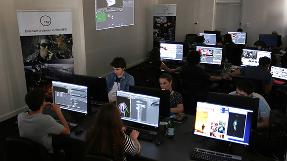 Students sit at desks using computer monitors in a dark classroom with a projector displaying software.