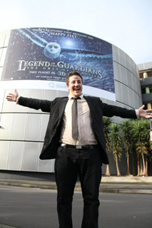 A man in a black suit and tie stands with arms wide open in front of a Legend of the Guardians movie poster.