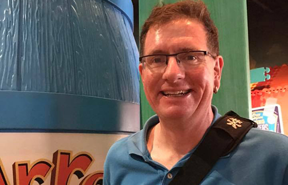 A man with glasses and a blue polo shirt smiles in front of a large blue textured structure.