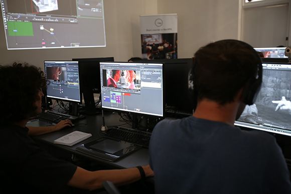 Two people sit at desks with multiple monitors displaying video editing software in a dark studio.