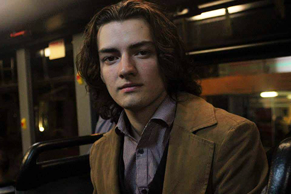A young man with wavy brown hair looks toward the camera while wearing a brown jacket.