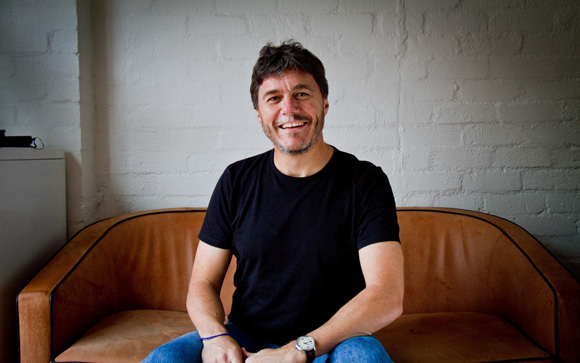 A smiling man with dark hair and a beard wearing a black t-shirt sits on a tan leather sofa.