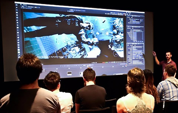 People sitting in a dark room watching a video editing software interface projected on a large screen.