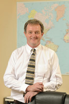 A man wearing a white dress shirt and patterned tie stands before a large world map.