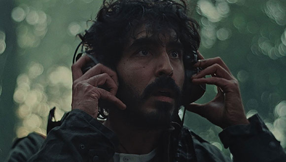 A man with dark curly hair and a mustache holds headphones to his ears against a blurred green background.