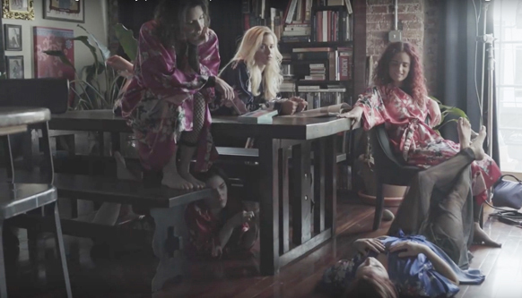 Four women wearing colorful robes lounge in a room with wooden furniture and bookshelves.