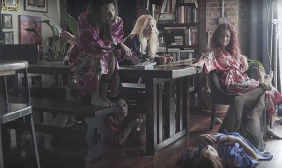 Five women in colorful robes lounge around a wooden table and on the floor in a room with bookshelves.