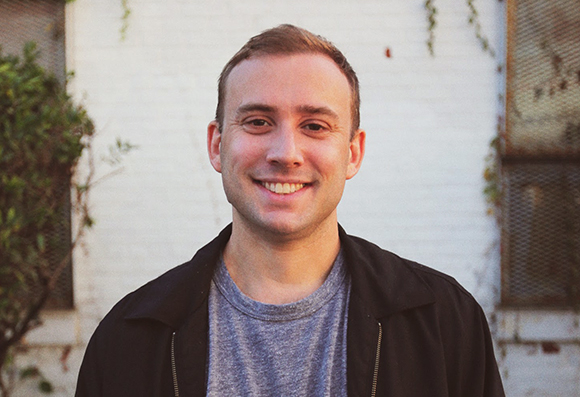 A smiling man with short brown hair wearing a black jacket and grey t-shirt against a white brick wall.