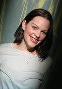A smiling woman with shoulder-length dark hair wearing a light-colored shirt and a thin necklace.