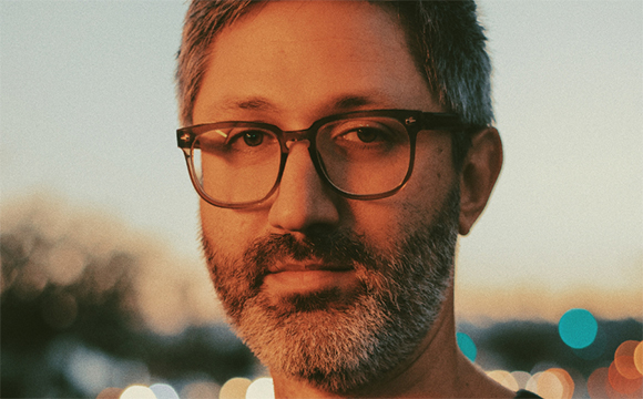 Close-up portrait of a man with short hair, glasses, and a salt-and-pepper beard against a sunset background.